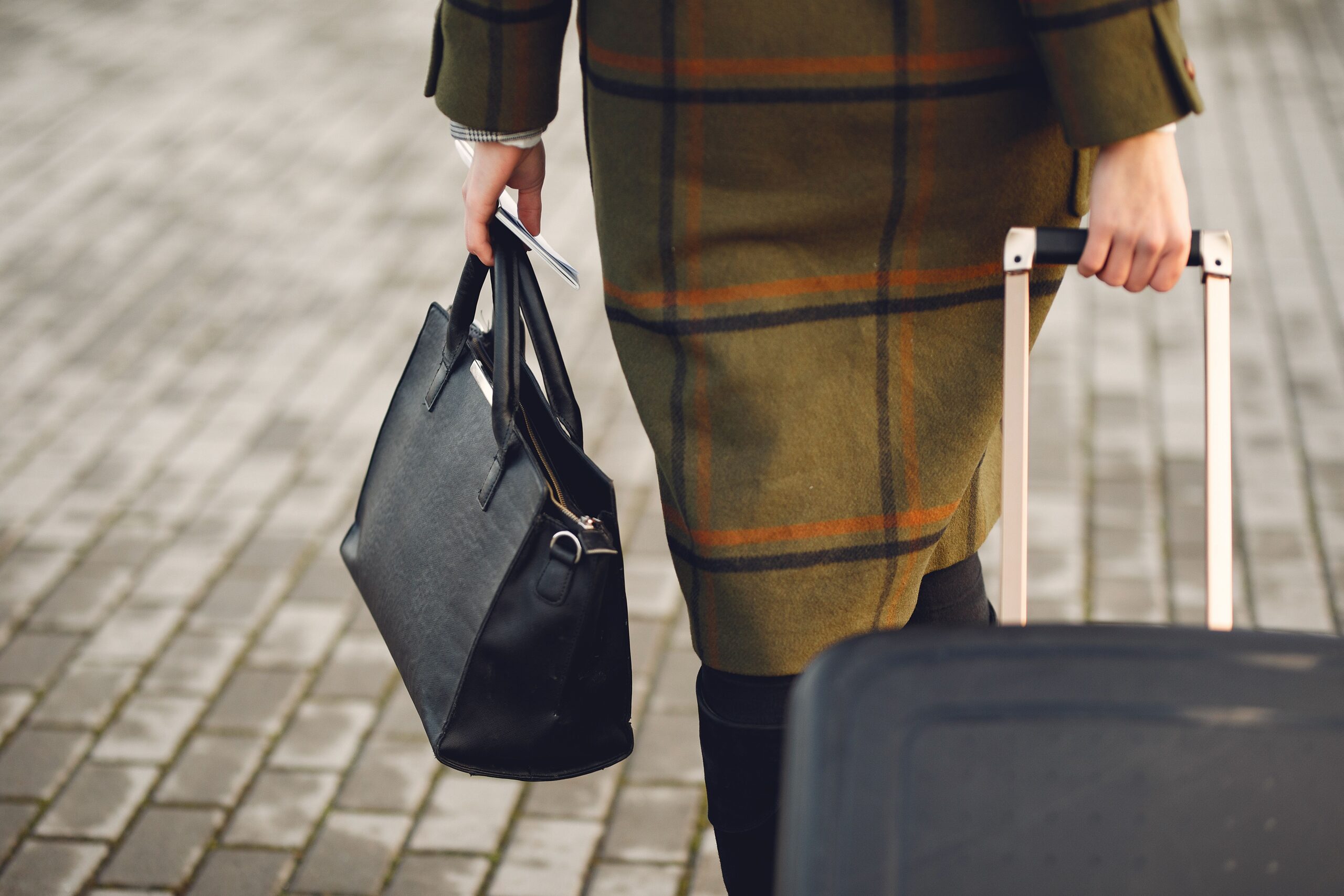 Home Back view of unrecognizable young female traveler in warm trendy plaid coat strolling on pavement with suitcase and bag while taking passport and tickets on city street in cold season